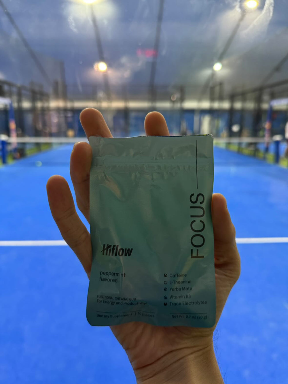 A person's hand holding a Hiflow Focus gum pouch inside a padel court.