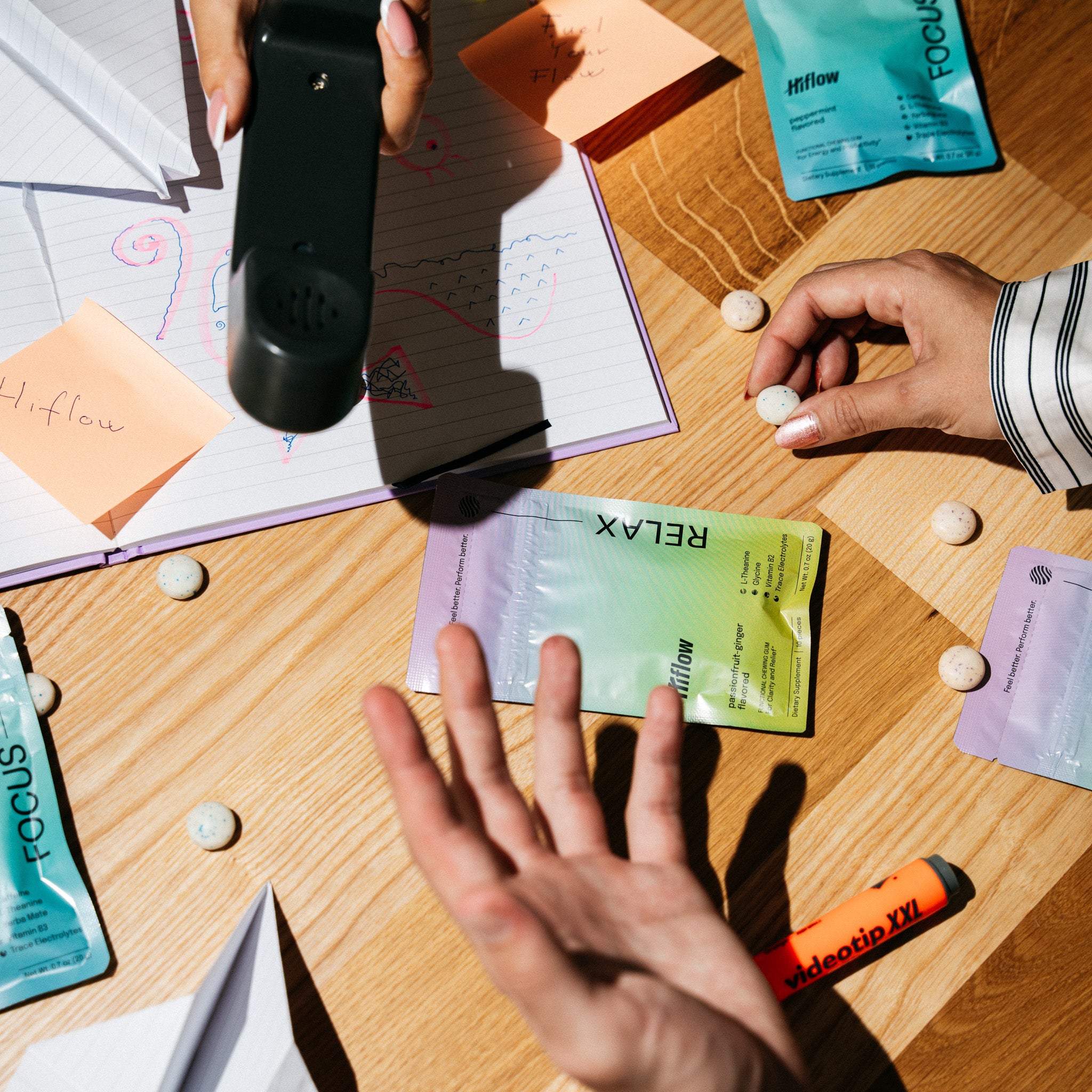 An image of a chaotic office desk with Hiflow gum pouches, gum pieces, a phone, and three hands of three different people.
